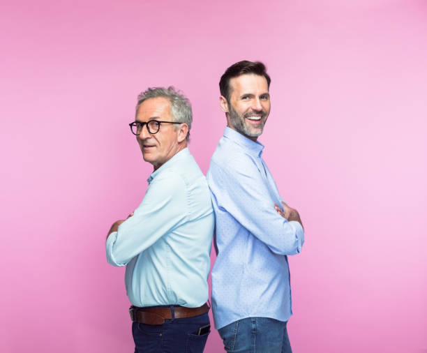 Happy father and son standing back to back Portrait of happy father and son standing back to back with arms crossed over pink background. three quarter length stock pictures, royalty-free photos & images