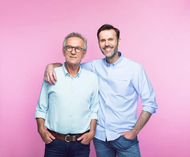 Smiling man with arm around senior father Portrait of smiling mid adult man standing with arm around senior father against pink background. arm around stock pictures, royalty-free photos & images