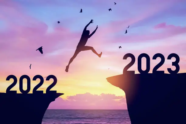 Photo of Silhouette man jumping between cliff with number 2022 to 2023 and birds flying at top of mountain. Freedom challenge and travel adventure holiday concept.