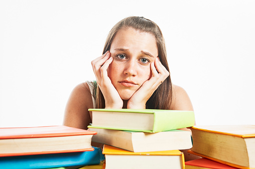 Female student looks mournfully at the camera as she sits at her desk loaded with hardcover textbooks.