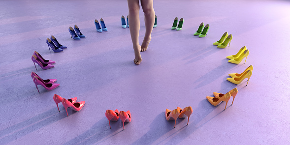 A woman with bare feet and legs standing on tiptoes in the centre of a circle created from generic high heels stiletto shoes arranged in rainbow colour order against a plain lilac background.