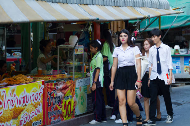 Thai university students near CRU university in Bangkok  Chatuchak are walking in street and are passing street food vendors and kitchens. One of students in foreground is a transgender. Younger students are buying food in background
