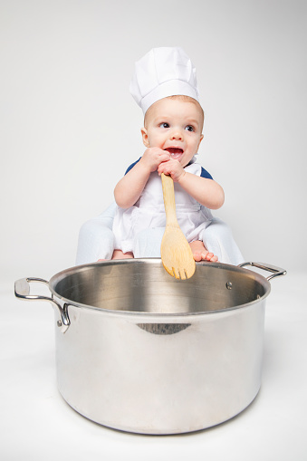 A 6 month old baby boy with a chef hat and apron holding a wooden spoon with a big pot nearby.