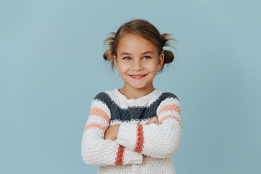 Confident smiling little girl in striped sweater standing with hands crossed over blue background.