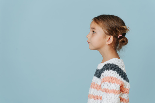 Side view of a cute little girl in striped sweater, her short hair in buns over blue background.