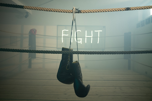 Boxing gloves hanging on a rope on empty boxing ring for training in dark gym. Neon fight sign is on the wall by the ring.