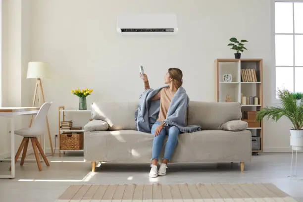 Woman who's sitting on sofa under warm plaid in living room switches off her air conditioner on wall. Young girl adjusting modern AC system, regulating temperature and enjoying cool fresh air at home