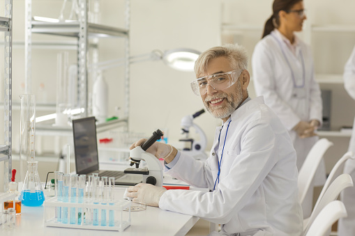 Portrait of senior male scientist working with microscope, laptop and lab equipment. Grey-haired researcher chemist sitting at desk looking at camera at modern scientific medical laboratory