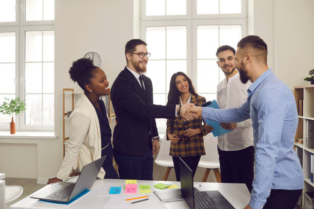 Male leader of a multiracial business team greets a new team member shaking his hand. People in a business meeting. Male leader of a multiracial business team greets a new team member shaking his hand. Concept of business relationships, hiring and promotion. new hire stock pictures, royalty-free photos & images