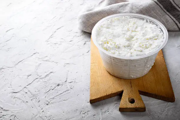 Photo of Ricotta cheese and wooden cutting board on white plaster background. Space for text.