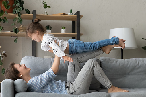 Happy mom lifting little daughter kid up in air, playing airplane with girl, lying on couch, keeping balance, doing acroyoga base with child, enjoying motherhood, active games, exercising