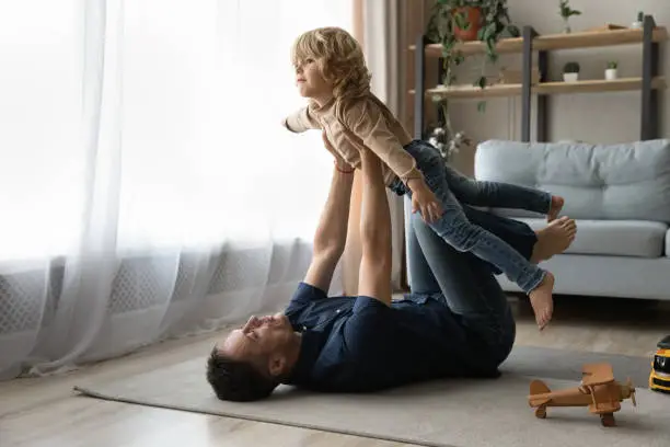Focused strong daddy playing active sportive games with preschool son, lying on heating carpeted floor with toys, holding boy with flying hands for making airplane, doing acroyoga base