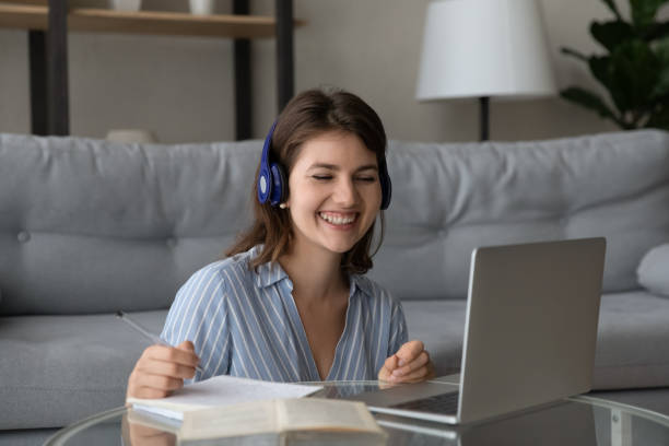 Cheerful student girl in wireless earphones making video call Cheerful student girl in wireless earphones making video call to teacher, watching learning webinar on laptop, studying foreign language on online class, writing audio lesson notes, laughing, online tutors stock pictures, royalty-free photos & images