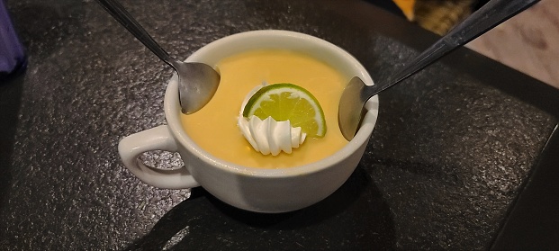 Delicious Key Lime Pie, homemade and shown uniquely in a large mug with 2 spoons, key lime wedge and whip cream dotted on the top.