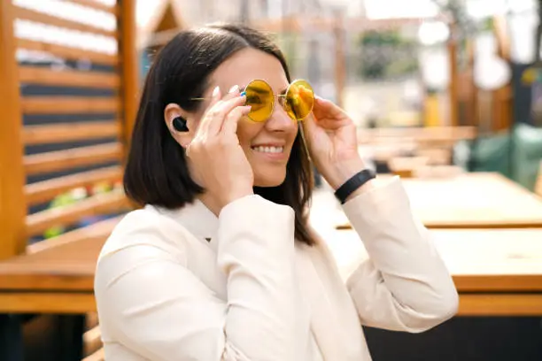 A happy business woman with mini in-ear headphones puts on yellow sunglasses. A sunny summer day in the city.