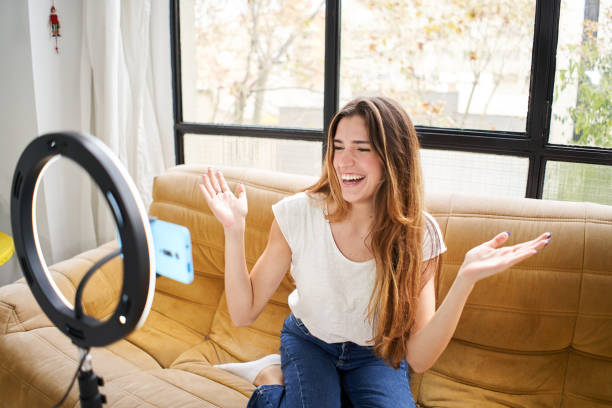 Modern influencer. Female vlogger making social media video while sitting indoors sofa home shooting with mobile smart phone camera.