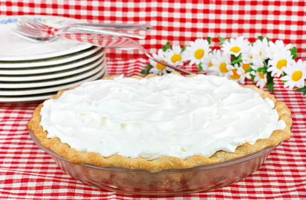 One whole whipped cream pie on a red and white tablecloth with flowers and plates in the background.  Could be any type of cream pie.
