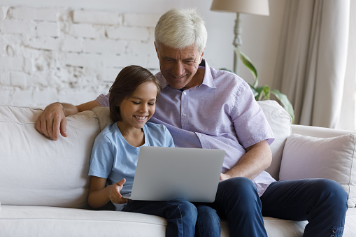 Happy gen Z grandkid boy and mature granddad watching movie on laptop at home, siting on sofa in living room, using computer, browsing internet, using online app for shopping, making video call