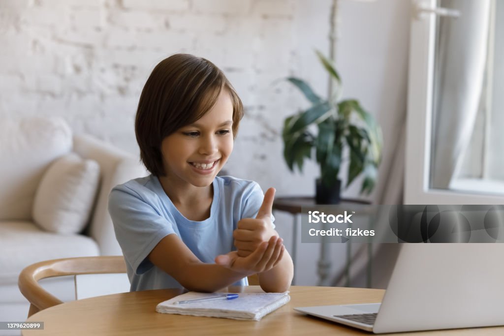 Smiling boy with hearing disability studying online from home Smiling positive teen pupil boy with hearing disability studying online from home, talking on video on laptop, speaking, using sign deaf language, showing gestures at screen. Childhood, education Deafness Stock Photo