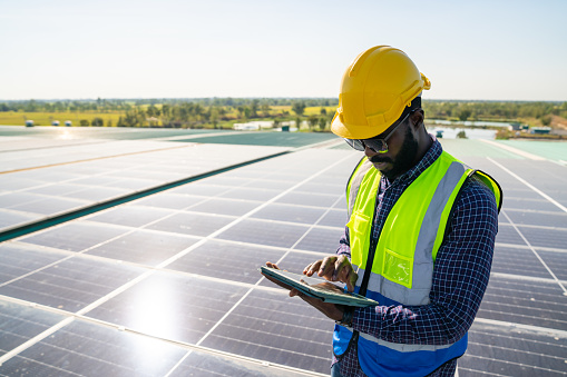 Professional African man engineer using digital tablet maintaining solar cell panels on building rooftop. Male technician working outdoor on ecological solar farm construction. Renewable clean energy technology concept