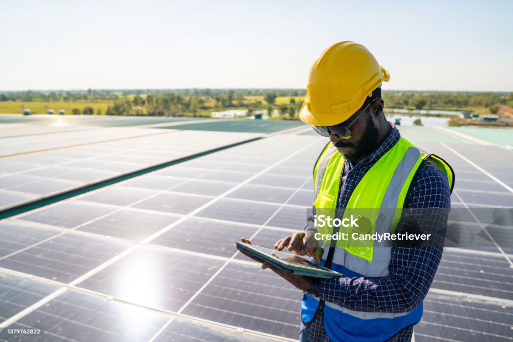 African man engineer using digital tablet maintaining solar cell panels on building rooftop. Professional African man engineer using digital tablet maintaining solar cell panels on building rooftop. Male technician working outdoor on ecological solar farm construction. Renewable clean energy technology concept Solar Panel Stock Photo