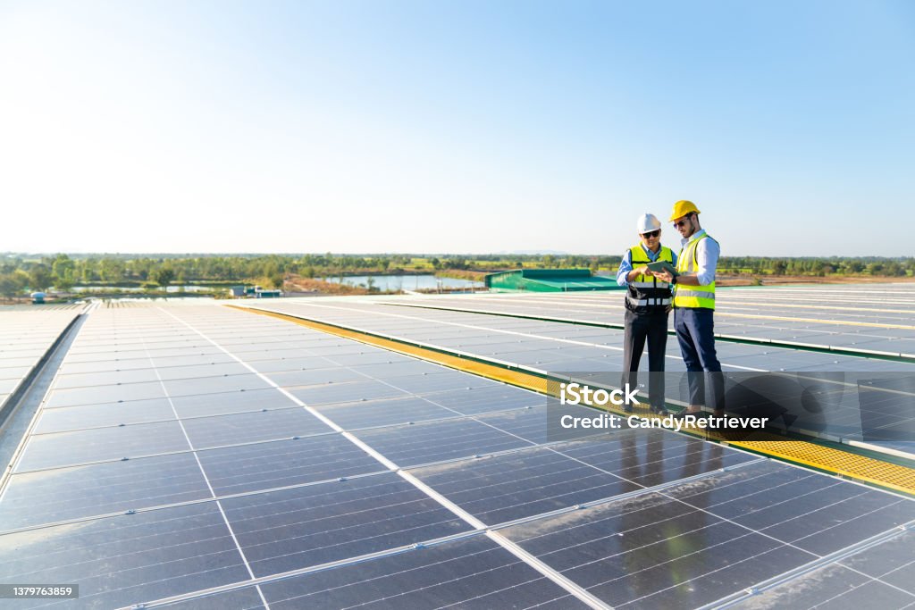 Professional man engineer using digital tablet maintaining solar cell panels together on building rooftop. Professional man engineer using digital tablet maintaining solar cell panels together on building rooftop. Technician team working on ecological solar farm. Renewable clean energy technology concept Solar Power Station Stock Photo