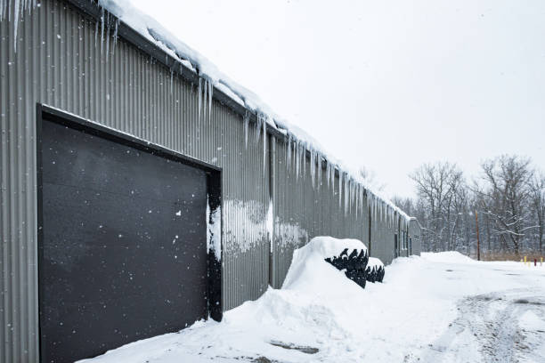 Winter snow storm icicles hanging from the edge of the roof of an industrial factory or warehouse facing a large, frozen asphalt parking lot.