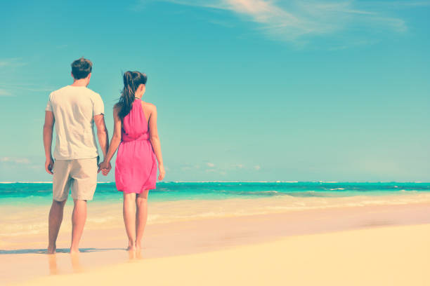 Couple standing on beach travel holding hands. Rear view of multiethnic romantic couple on shore. Young partners are watching sea. Tourists are enjoying their summer vacation at beach.