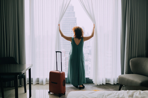 An unrecognizable woman drawing the curtains in a hotel room upon arrival to enjoy the view.