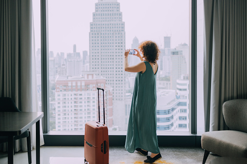 An anonymous woman using her mobile phone to take a picture of her hotel room window view of the city.