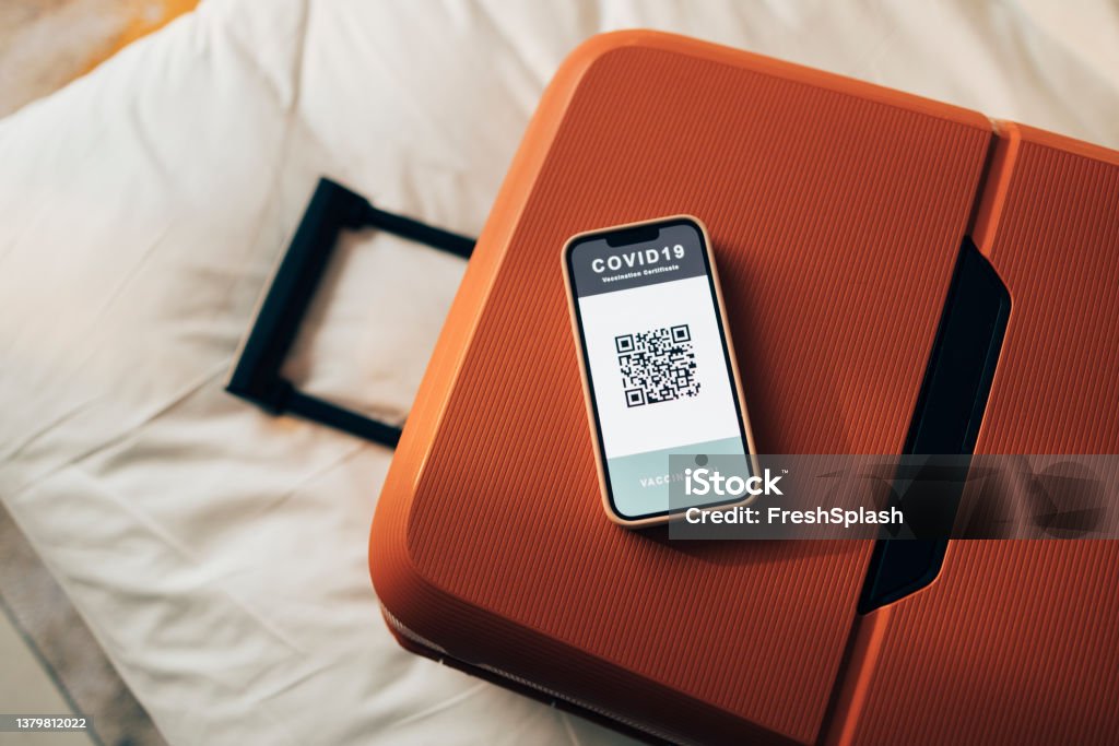 Getting Ready for Vacation: A Red Suitcase with a Smartphone Screen Showing a Covid Vaccination QR Code A packed suitcase and a mobile phone showing a digital covid passport QR code on a bed with white sheets. Suitcase Stock Photo