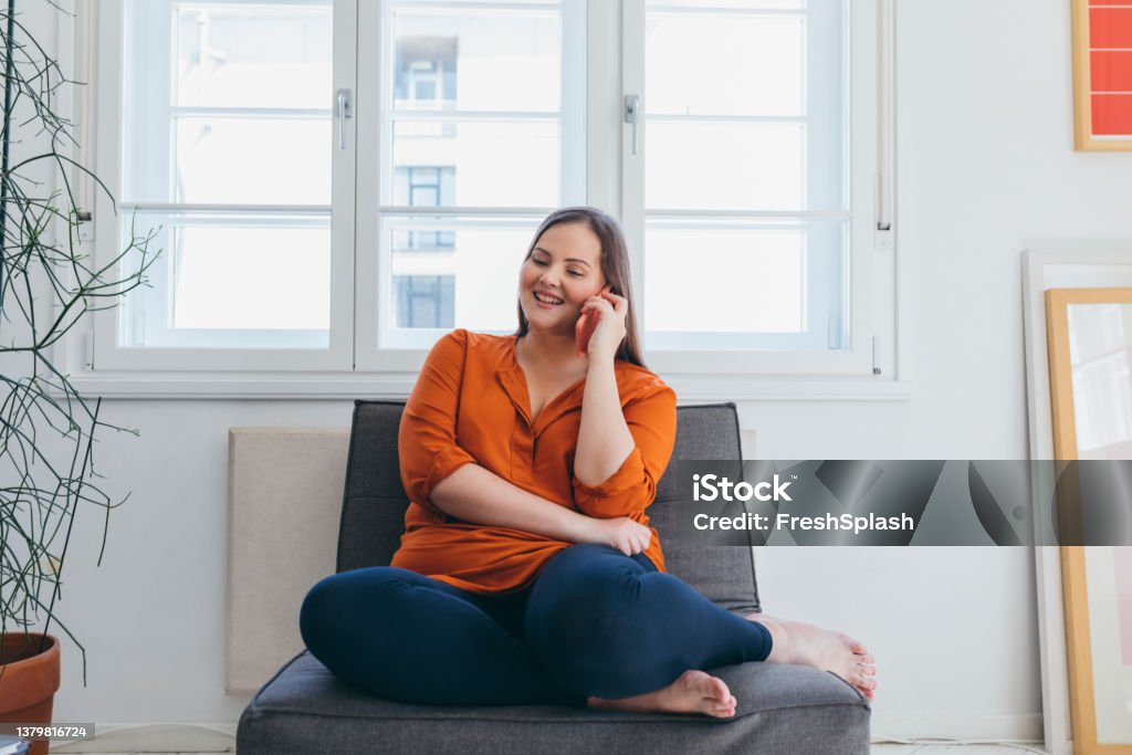 Smiling Plus Size Woman Wearing an Orange Shirt Talking on the Phone while Sitting in the Living Room A happy overweight woman talking on the phone while sitting on an armchair in the living room. Obesity Stock Photo