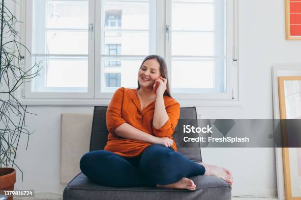 Smiling Plus Size Woman Wearing An Orange Shirt Talking On The Phone While Sitting In The Living Room Stock Photo - Download Image Now