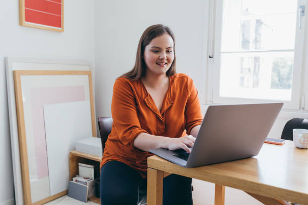 femme souriante de taille plus assise à la table de son bureau à domicile et tapant quelque chose sur son ordinateur portable - heavy work photos et images de collection