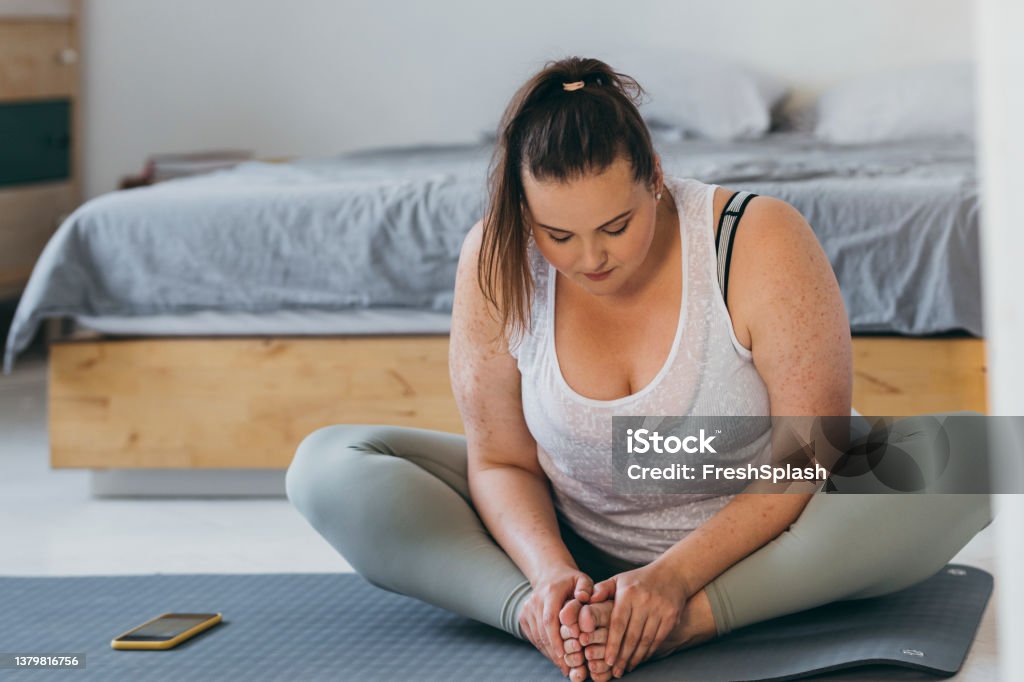 Beautiful Plus Size Woman Doing Yoga in her Bedroom in the Morning A young overweight woman doing stretching exercises in her bedroom. Obesity Stock Photo