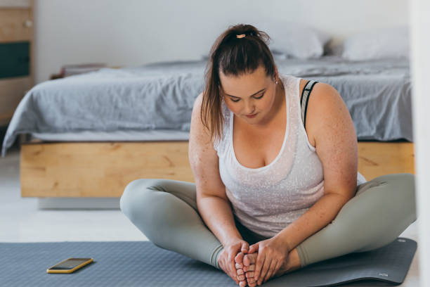 Beautiful Plus Size Woman Doing Yoga in her Bedroom in the Morning A young overweight woman doing stretching exercises in her bedroom. obesity stock pictures, royalty-free photos & images