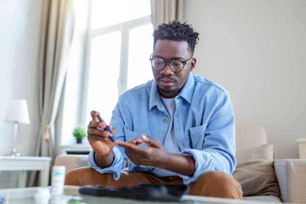 african man is sitting at the sofa and taking blood from his finger due to diabetes. the daily life of a man of african-american ethnicity person with a chronic illness who is using glucose tester. - blodsockerprov bildbanksfoton och bilder