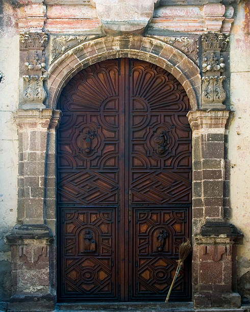 A very large carved wooden door is the entrance to an old colonial church in San Miguel de Allende Mexico. A staw broom is standing next to the entrance.