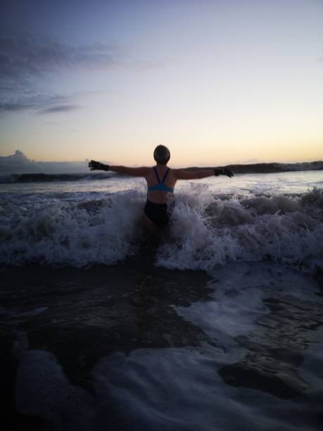Open Water Swimming at Sunset Burry Port, Wales, UK: April 03, 2022: A women jumps through the waves while entering the sea on a cold water ocean swim at dusk. woman cold water swimming stock pictures, royalty-free photos & images