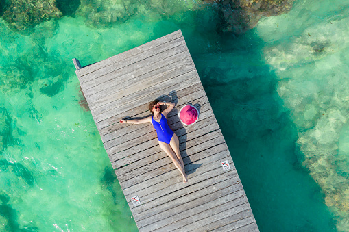Aerial view of woman laying on wooden pier at sunny summer day in Cancun, Mexico, top view. Young sexy woman wearing bright swimsuit in summertime in Caribbean