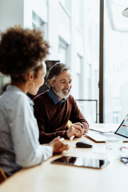 uśmiechnięty doradca finansowy i biznesmen rozmawiający z koleżanką z mieszanej rasy w biurze - leadership business women senior adult zdjęcia i obrazy z banku zdjęć