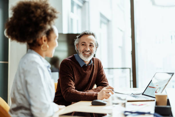 uśmiechnięty doradca finansowy i biznesmen rozmawiający z koleżanką z mieszanej rasy w biurze - business meeting teamwork meeting mature adult zdjęcia i obrazy z banku zdjęć