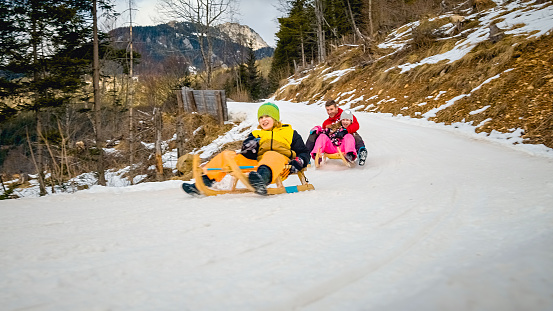 Happy Caucasian family, father, and two children sliding on wooden sleds along a snowy mountain road.