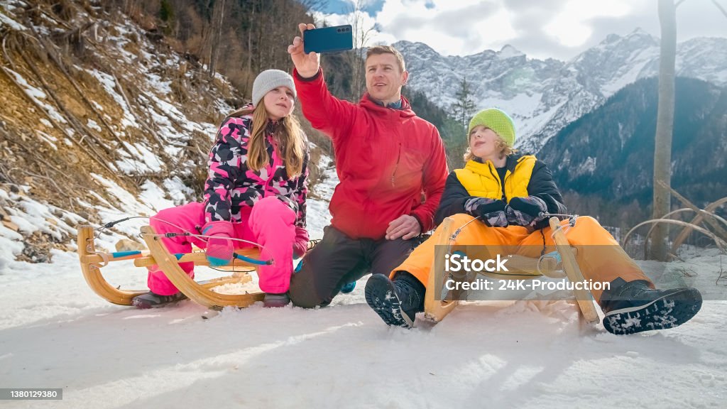 Father with children taking selfies while sitting on sleds Father with children taking selfies while sitting on wooden sleds, with a beautiful mountain landscape in the background. 10-11 Years Stock Photo