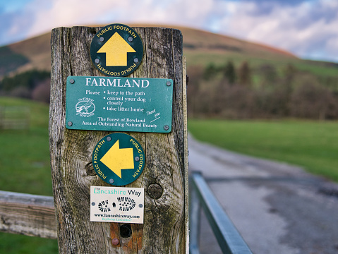 Green circular waymarkers fixed to a weathered wooden post near Dunsop Bridge in Lancashire, UK. A green rectangular sign reminds walkers to respect the farmland.