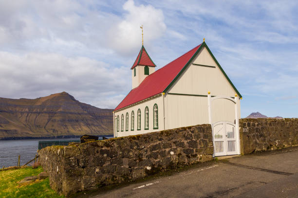 Kunou, Faroe Islands - 02 May 2018: Kunoy Church on a hill. Steep coast of the island of Kalsoy in the background.