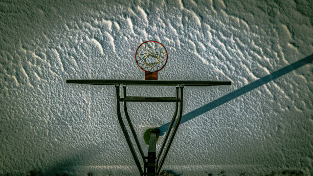 Low-angle view of a worn red basketball hoop with a frayed net against a pale cloudy sky, conveying urban grit, outdoor recreation, nostalgia and the silence of an empty court.
