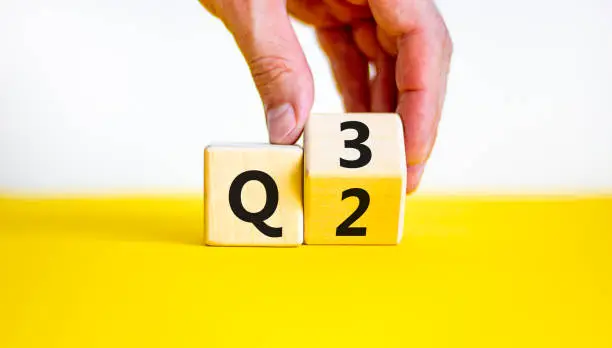 Photo of From 2nd to 3rd quarter symbol. Businessman turns a wooden cube and changes words 'Q2' to 'Q3'. Beautiful yellow table, white background. Business, happy 3rd quarter Q3 concept, copy space.