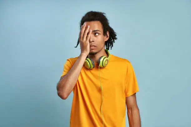 Bored African young man covers the face, sighs from unpleasure, made bad mistake, wears orange outfit, has stereo headphones, stands against blue wall, blank space for your advertising content