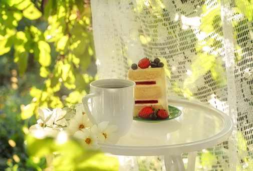 Beautiful porcelain coffee cup and slice of cake on glass table decorated with white flowers in summer garden in sunlight. Copy space. Summer drinks concept.